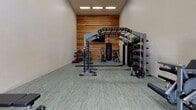 This dedicated fitness center is thoughtfully designed to support a full range of workout routines in a clean, modern environment. The space features a warm wood-plank accent wall that adds a natural, inviting aesthetic and serves as a striking backdrop throughout the room. The flooring consists of durable, light-toned gym tiles that provide both cushioning and easy maintenance.

The cardio zone is well-appointed with two treadmills positioned side by side beneath a wall-mounted flat-screen television, making for an engaging workout experience. An elliptical trainer and a recumbent exercise bike round out the cardio offerings, catering to a variety of fitness preferences.

The strength training area is a standout feature, anchored by a large multi-functional rig with overhead bars, suspension training attachments, a punching bag, and weight storage. A cable machine and a dedicated dumbbell rack with an adjustable bench provide additional options for resistance training. A Torros functional trainer further expands the versatility of the space.

The room benefits from notably high ceilings, which contribute to an open and airy atmosphere. A set of glass-paned double doors at one end of the space allows natural light to filter in and provides a connection to the outdoors. Recessed overhead lighting ensures the space is well-lit at all times, creating a bright and motivating workout environment.