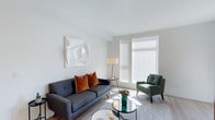 This single-floor space showcases a thoughtfully designed living area that balances contemporary aesthetics with functional comfort. The room is characterized by crisp white walls and smooth ceilings that amplify the abundance of natural light streaming through large, floor-to-ceiling windows dressed with horizontal blinds, allowing for adjustable privacy and light control.

The living area is anchored by a charcoal-gray mid-century modern sofa adorned with warm rust-orange velvet throw pillows, complemented by a forest green accent chair that introduces a rich color contrast. A round glass-top coffee table sits atop a light-toned patterned area rug, adding a sense of openness to the space. A slim floor lamp provides supplementary ambient lighting alongside a small side table, enhancing the room's layered lighting design.

Wall décor includes a striking white geometric sculptural canvas, adding texture and visual depth without disrupting the minimalist color scheme. The light hardwood flooring ties the space together, contributing to the overall airy and modern ambiance. The layout feels well-proportioned, offering a comfortable and inviting environment suited for relaxed living.