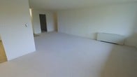 This single-floor space presents a straightforward, functional layout centered around a generously sized open main living area. The room is finished with wall-to-wall light gray carpeting that extends throughout, providing a clean and uniform foundation underfoot. The walls are painted in a soft, neutral off-white tone, contributing to a bright and airy atmosphere despite the absence of visible windows in the main area. A baseboard heating unit is positioned along one wall, indicating an efficient heating solution integrated into the space. The open floor plan allows for flexible furniture arrangement, accommodating a variety of living configurations. A doorway visible toward the rear of the main room leads to an additional space, suggesting further rooms or utility areas beyond the primary living zone. The overall design is minimal and practical, with smooth ceilings and clean wall lines that emphasize the sense of openness. The neutral palette and open layout make this space highly adaptable, offering a versatile foundation suitable for a range of furnishing styles and functional needs.