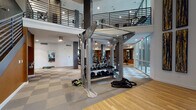 This two-story fitness facility, branded "Elevation," is thoughtfully designed to support a full range of workout experiences. The main level opens into a welcoming lounge area finished with warm hardwood flooring, a wall-mounted television, built-in wood shelving, and modular hexagonal seating in yellow, grey, and charcoal tones. Soaring floor-to-ceiling windows flood the space with natural light and offer views of an outdoor pool and palm trees, enhancing the open, airy ambiance. The motivational "Rise Above" Elevation logo adorns the lounge wall, setting an energetic tone throughout. The fitness floor features a checkerboard-pattern carpet tile layout and is divided into distinct training zones. A central strength area houses a freestanding multi-functional rig, a full dumbbell rack, a Smith machine, adjustable benches, and battle ropes for functional training. A dedicated cardio zone is equipped with treadmills, ellipticals, stair climbers, stationary bikes, and rowing machines, all positioned near mirrored walls with warm-toned wood accents. A separate functional training room includes plyo boxes, kettlebells, and resistance equipment against a deep navy accent wall. An upper mezzanine level, accessible via an interior staircase with metal railings, provides additional equipment and overlooks the main floor below.