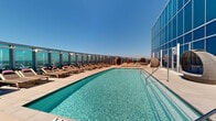 Perched high above Long Beach at 777 E Ocean Blvd, this expansive rooftop amenity deck offers a resort-caliber outdoor experience with sweeping panoramic views of the city skyline, coastline, and surrounding landscape. The centerpiece of the space is a generously sized lap-style swimming pool with crystal-clear blue water, flanked by a long row of wood-framed lounge chairs positioned to maximize sun exposure and the breathtaking vistas beyond the glass perimeter railing. Adjacent to the pool, a bubbling spa provides a more intimate relaxation option, accompanied by cozy pod-style lounge seating nestled against the striking blue glass curtain wall of the building. The outdoor kitchen area is fully equipped with a professional-grade stainless steel grill and prep surfaces, making it ideal for entertaining. Intimate dining nooks with bistro tables and chairs are positioned along the glass railing, offering front-row seats to the expansive views. The deck is finished in large-format neutral stone pavers throughout, lending a clean, modern aesthetic. The floor-to-ceiling blue glass facade of the building serves as a dramatic architectural backdrop, while the open-air design ensures abundant natural light and fresh ocean breezes throughout the entire space.