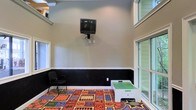 This single-floor space is designed as a functional activity or common room, offering a welcoming environment suited for leisure and entertainment. The room features a two-tone wall treatment, with light gray on the upper half and dark paneling on the lower half, creating a clean and defined visual contrast throughout the space.

A wall-mounted television is positioned at an elevated height, making it easily viewable from anywhere in the room. Seating is provided by a simple task chair, while a white activity table with a green surface sits atop a vibrant, colorful carpet featuring bold patterns of numbers, letters, and classic board game motifs — adding an engaging and playful element to the space.

Large multi-pane windows line one side of the room, allowing generous amounts of natural light to filter in and providing views of the surrounding exterior. The high ceiling enhances the sense of openness, and the elevated window placement near the top of the walls further contributes to the airy feel of the interior.

Overall, this space presents a casual and functional layout well-suited for shared activities, whether for recreation, entertainment, or group use. The combination of practical furnishings, bold floor coverings, and ample natural light creates a bright and inviting atmosphere.