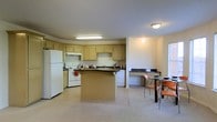 This two-floor residential space is thoughtfully laid out to support comfortable and functional everyday living. The kitchen serves as a central hub, featuring light wood cabinetry that extends to the upper walls, offering generous storage throughout. A dark-countered peninsula with an integrated sink provides additional workspace and separates the kitchen from the adjacent areas, while a freestanding central island adds further prep and casual gathering space. Standard appliances — including a white refrigerator, electric range, and dishwasher — are neatly integrated into the layout. Overhead fluorescent and flush-mount lighting keep the kitchen bright and practical. Adjacent to the kitchen, a built-in desk area with dark countertop provides a convenient workspace or study nook. The dining area nearby accommodates a table and chairs, making it well-suited for meals and everyday use. Light-toned walls and neutral flooring throughout create a clean, cohesive aesthetic that runs across both floors. The open flow between the kitchen, workspace, and dining zones gives the main level an interconnected, spacious feel that supports both daily routines and casual entertaining.