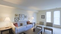 This single-floor residential space offers a warm and welcoming atmosphere defined by a clean, neutral color palette and thoughtfully arranged living areas. The living room serves as the heart of the space, featuring light-toned walls that enhance the sense of openness and allow natural light to fill the room. A plush sofa anchored by decorative throw pillows sits across from a pair of patterned accent chairs, creating a comfortable and functional seating arrangement centered around a dark wood coffee table. A side table with a classic lamp adds a soft, ambient glow to the space. The walls are tastefully decorated with a curated gallery of framed artwork and decorative accents, lending the room a lived-in yet polished character. Light-colored carpeting runs underfoot, contributing to the room's overall warmth and comfort. A window on the far wall allows natural light to filter in, brightening the space and enhancing its airy feel. A doorway visible in the background suggests additional rooms and smooth flow throughout the layout. The overall ambiance is calm, tidy, and welcoming, making it a practical and comfortable place to call home.