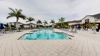 Located at 9940 Estero Oaks Drive in Fort Myers, Florida, this outdoor community amenity space offers a resort-style experience designed for relaxation and recreation. The centerpiece is a generously sized swimming pool with clearly marked lap lanes, accommodating both leisure swimmers and those seeking structured exercise. The pool features a graduated entry point with handrail access, making it accessible and welcoming for all users.

Surrounding the pool is an expansive paver deck finished in light-toned stone tile, providing ample space for sunbathing and socializing. Numerous lounge chairs and seating clusters are arranged throughout the deck, offering flexible options for poolside relaxation. Brightly colored Adirondack chairs are positioned along the lawn area, adding a casual, inviting touch to the setting.

Tall, mature palm trees are thoughtfully distributed across the space, providing natural shade and lending the area a distinctly tropical Florida character. Manicured landscaping with lush greenery and flowering shrubs frames the perimeter, enhancing the visual appeal.

A well-appointed clubhouse building is situated adjacent to the pool area, featuring a covered pergola and large glass doors that connect indoor and outdoor spaces seamlessly. A scenic water view is visible in the background, with the broader residential community providing an attractive backdrop. The overall ambiance is open, airy, and evocative of a luxurious Florida lifestyle.
