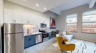 Located in the heart of downtown Cincinnati at 135 W Fourth St, this thoughtfully designed urban residence offers a compelling blend of modern finishes and city-inspired living. The open-concept layout seamlessly connects the kitchen and living area, creating an inviting, functional space filled with natural light from large windows that frame striking views of Cincinnati's historic architecture and skyline.

The kitchen is a standout feature, outfitted with two-tone cabinetry — crisp white upper cabinets paired with slate blue lower cabinets — complemented by a white subway tile backsplash, light countertops, and a full suite of stainless steel appliances including a refrigerator, dishwasher, electric range, and over-the-range microwave. The adjacent living area benefits from elevated ceilings with exposed beams and provides a comfortable, well-lit setting with direct city views.

The bedroom is a calm, neutral retreat with soft carpet flooring, generous wall space, and a relaxed, restful ambiance. The bathroom features a fully tiled walk-in shower with a clean white subway tile pattern in a herringbone-style layout and matte black fixtures, delivering a polished, contemporary aesthetic throughout.