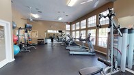 This fitness center is a spacious, single-floor facility designed to support a comprehensive workout experience. The room features soaring ceilings that create an open, airy atmosphere, enhanced by a row of large, multi-pane windows along one wall that flood the space with abundant natural light and provide views of the outdoors. Ceiling fans are mounted overhead to maintain air circulation throughout the room.

The cardio section is well-appointed with a lineup of True-brand treadmills and elliptical machines, offering ample equipment for aerobic training. A dedicated free weights area includes a full rack of dumbbells in varying sizes, medicine balls, and a weight bench, catering to strength training needs. Full-length mirrors along one wall visually expand the space and allow users to monitor their form during exercise.

Multiple wall-mounted flat-screen televisions are positioned throughout the room for entertainment during workouts. The flooring consists of durable, dark rubber matting, providing both safety and cushioning. The warm-toned walls and natural light contribute to an energizing yet comfortable environment suited for a wide range of fitness activities.