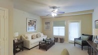 This single-floor space offers a comfortable and well-appointed living environment designed with both style and functionality in mind. The living room serves as the heart of the space, featuring warm, neutral-toned walls that create an inviting and relaxing atmosphere. Plush wall-to-wall carpeting adds a sense of warmth underfoot, while a classic multi-blade ceiling fan with integrated lighting provides both airflow and ambient illumination. Natural light flows generously into the room through multiple windows dressed with clean white blinds and a soft Roman shade, enhancing the bright and airy feel of the interior. A glass-paneled door provides direct access to an outdoor area, further extending the sense of openness. The living room is thoughtfully laid out, offering ample space for seating and a central coffee table, making it well-suited for both relaxing and entertaining. Decorative framed artwork adorns the walls, adding personality and visual interest to the space. The overall ambiance is calm, clean, and cohesive, with a neutral color palette that lends itself well to a variety of furnishing styles.