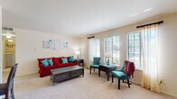 This single-floor residence offers a thoughtfully arranged layout that maximizes comfort and functionality. The living room serves as the heart of the space, featuring plush carpeting, neutral walls, and a bank of multiple windows fitted with blinds and sheer curtains that invite generous natural light and outdoor views. The room provides ample space for both a sofa arrangement and a dedicated seating area, making it versatile for both relaxing and entertaining.

The galley-style kitchen runs along a narrow corridor, lined on both sides with cream-toned cabinetry offering substantial storage, a stainless steel sink, a dishwasher, and a white range and refrigerator. Tile flooring throughout the kitchen provides easy maintenance. Adjacent to the kitchen, a dedicated dining area accommodates a full-sized table and chairs beneath a multi-bulb chandelier, with the kitchen appliances conveniently nearby.

The bedroom is a calm, carpeted retreat with a window framing outdoor greenery, neutral walls, and built-in wall shelving. The full bathroom features a single-sink vanity with cabinet storage below, a mirror, tile flooring, and a shower-tub combination accented by a colorful curtain. Throughout, the space maintains a consistent, warm aesthetic with light tones and functional design.