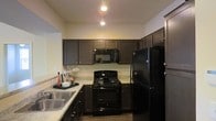 The kitchen serves as a central highlight of this single-story space, showcasing a cohesive and functional design. Dark espresso-toned cabinetry lines the walls, extending to the upper sections and providing generous storage throughout. The light-toned stone countertops offer a striking contrast against the darker elements, creating visual balance and a clean aesthetic. Black appliances, including a gas range, built-in microwave, and a full-size refrigerator, maintain a uniform and modern look throughout the kitchen. A double-basin stainless steel sink is positioned along one wall, complementing the overall palette. Pendant lighting and recessed ceiling fixtures illuminate the workspace efficiently, enhancing the warm ambiance of the room. The layout follows a practical U-shaped configuration, maximizing counter space and ease of movement. The flooring transitions naturally underfoot, and the open sightlines suggest a connection to adjacent living areas, contributing to an overall sense of openness and flow within the single-floor layout.