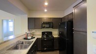 The kitchen serves as a central highlight of this single-story space, showcasing a cohesive and functional design. Dark espresso-toned cabinetry lines the walls, extending to the upper sections and providing generous storage throughout. The light-toned stone countertops offer a striking contrast against the darker elements, creating visual balance and a clean aesthetic. Black appliances, including a gas range, built-in microwave, and a full-size refrigerator, maintain a uniform and modern look throughout the kitchen. A double-basin stainless steel sink is positioned along one wall, complementing the overall palette. Pendant lighting and recessed ceiling fixtures illuminate the workspace efficiently, enhancing the warm ambiance of the room. The layout follows a practical U-shaped configuration, maximizing counter space and ease of movement. The flooring transitions naturally underfoot, and the open sightlines suggest a connection to adjacent living areas, contributing to an overall sense of openness and flow within the single-floor layout.