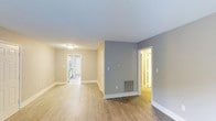 Located at 8501 New Brunswick Ln in Raleigh, North Carolina, this single-floor home offers a well-designed layout with a bright, neutral aesthetic throughout. The interior showcases light hardwood-style flooring that runs seamlessly across the main living areas, creating a cohesive and inviting atmosphere. Walls are finished in soft, warm neutral tones complemented by crisp white trim and baseboards, lending the space a clean and modern feel. The open main living area is generously proportioned, allowing for flexible furniture arrangements and easy flow between spaces. A floor vent indicates a central HVAC system for year-round climate comfort. An accent wall in a cool gray tone adds a subtle visual contrast to the warmer surrounding walls, providing a tasteful design detail. Doorways lead to additional rooms, including what appears to be a laundry area and other private spaces, all benefiting from the same consistent flooring and bright, neutral finishes. Flush-mount ceiling lighting provides even illumination throughout. The overall ambiance is clean, move-in ready, and versatile, well-suited for a variety of living arrangements. The single-level layout ensures ease of accessibility and navigation throughout the entire home.