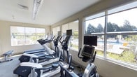 This single-level fitness center is designed to provide a bright, motivating environment for exercise. The space is lined with large windows fitted with roller shades, allowing generous amounts of natural light to fill the room while offering views of an outdoor pool area and mature trees beyond — creating a refreshing, open atmosphere during workouts.

The cardio equipment area is well-appointed with a row of treadmills, elliptical trainers, and stationary bikes, each unit equipped with individual display screens for entertainment or workout tracking. The machines are arranged to take full advantage of the window views, allowing users to look outward while exercising. The layout makes efficient use of the available floor space while maintaining clear pathways between equipment.

The room features neutral-toned walls and a low ceiling with flush-mounted lighting that complements the natural light from the windows. The overall aesthetic is clean and contemporary, with a practical, no-frills design focused on functionality. The fitness center's connection to the outdoors through its expansive windows gives the space an airy, energizing quality well-suited for an active lifestyle.