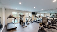 This single-floor amenity facility is thoughtfully designed to support both fitness and professional needs. The centerpiece is a generously sized fitness room outfitted with a comprehensive selection of cardio equipment, including treadmills, elliptical trainers, upright and recumbent stationary bikes, and a rowing machine. A full-length wall mirror amplifies the sense of space and allows users to monitor their form, while a wall-mounted flat-screen television provides entertainment during workouts. Recessed lighting illuminates the room evenly, and a row of large windows along one wall floods the space with natural light and offers views of an outdoor pool area and surrounding greenery, creating an energizing atmosphere. An additional fitness alcove provides further equipment space with its own mirrored wall. The facility also includes a sleek, modern meeting room featuring light wood-look tile flooring, recessed lighting, two conference tables with contemporary task chairs, a wall-mounted display screen, and elegant floor-to-ceiling drape panels that lend a polished, professional ambiance. A mezzanine-level walkway with dark metal railings overlooks a glass-walled racquetball court below, rounding out this versatile and well-appointed facility.