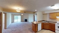 This single-floor residence is designed with an open-concept layout that seamlessly connects the kitchen and living areas, creating a spacious and functional environment. The kitchen features warm wood cabinetry, a dark laminate countertop, a corner sink, and a peninsula with additional storage and workspace. A white dishwasher is integrated into the cabinetry, and tile flooring grounds the kitchen space.

The kitchen flows directly into the expansive carpeted living area, which is bathed in natural light from a large sliding glass door that opens to an outdoor view of mature trees. A ceiling fan is positioned centrally over the living space, while a pendant light fixture illuminates the kitchen transition area. A textured ceiling adds subtle character throughout.

A gas fireplace serves as a focal point along one wall of the living area, adding warmth and visual interest. Recessed lighting and HVAC vents are integrated into the ceiling. A hallway visible from the living space leads to additional rooms, hinting at a thoughtful and practical floor plan throughout the home.