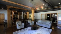 This single-floor space showcases a compelling fusion of industrial and traditional design elements. The open layout is defined by dark-painted ceilings with exposed ductwork and track lighting, which contrasts with warm-toned wooden structural columns and detailed millwork that anchor the room visually. The ceiling height adds a sense of volume and openness throughout the space.

The lounge area is furnished with a neutral-toned sofa and matching armchairs arranged around a dark oval coffee table, set atop a patterned area rug. This seating arrangement creates a comfortable, conversation-friendly focal point. Adjacent to the lounge, a dining or bar area features high-backed wooden chairs paired with tall tables, alongside a substantial wooden shelving unit displaying decorative items.

Along the right wall, two framed glass doors with wooden trim provide access to additional rooms or utilities, maintaining a cohesive aesthetic. Artwork on the walls and warm accent lighting contribute to an inviting yet sophisticated ambiance. The overall character of the space suits a variety of uses, from professional gatherings to social events.