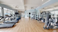 Located at 1001 Cox Grade Rd in Panama City Beach, FL, this single-floor fitness center is thoughtfully designed to provide a comprehensive workout experience in a bright, welcoming environment. The main exercise area features light hardwood-style flooring, coffered ceilings with ceiling fans, and recessed lighting that create an airy, open atmosphere. Soft blue walls and floor-to-ceiling windows line one side of the space, flooding the interior with natural light and offering views of the outdoor pool and surrounding community. The cardio zone is well-equipped with multiple treadmills, elliptical trainers, and stationary bikes, all positioned to take advantage of the expansive window views. The strength training section includes cable machines, weight benches, a multi-station rack, and a full dumbbell rack, complemented by large wall mirrors that enhance the sense of space. A separate studio room provides a versatile area for group fitness or individual training, outfitted with stability balls, TRX equipment, plyometric boxes, step platforms, and a wall-mounted TV. An additional flex area features a wall-mounted TV, a floating counter, and a water dispenser, serving as a convenient rest and refreshment station. Direct access to the outdoor area is available through glass-paned doors.
