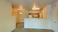This single-floor residential space offers a practical and functional open-concept layout that seamlessly connects the living area to the kitchen. The living space features neutral-toned wall-to-wall carpeting, creating a warm and comfortable atmosphere, with soft ambient lighting provided by a classic pendant light fixture overhead.

A half-wall partition defines the boundary between the living area and the kitchen, adding a sense of structure while maintaining an open feel throughout. The kitchen is well-appointed with warm wood cabinetry providing ample storage, and is equipped with white appliances including a refrigerator, microwave, and oven. Flush ceiling lighting ensures the kitchen is well-illuminated for everyday use.

The overall color palette is light and neutral, with white walls and trim throughout, contributing to a clean and airy ambiance. The layout is efficient and straightforward, making good use of the available space. A doorway visible from the kitchen suggests additional rooms or storage areas beyond, further enhancing the functional nature of this single-floor residence. The design prioritizes practicality while maintaining a comfortable and inviting living environment.