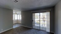 This single-story space centers around a spacious living area finished with plush carpet flooring and neutral gray walls that create a clean, understated backdrop. A textured popcorn ceiling adds a classic touch overhead, while a ceiling fan with a built-in light fixture provides both air circulation and ambient lighting. Natural light fills the room through multiple windows fitted with white horizontal blinds, as well as a large sliding glass door framed in a grid-pane design. The sliding door opens to an outdoor area and frames views of mature trees and lush greenery beyond, giving the interior a pleasant connection to the outdoors. Floor-level HVAC vents are positioned throughout the space, indicating a ducted climate control system. White baseboards provide a crisp contrast against the gray walls and carpeted floors. The overall ambiance is calm and neutral, offering a versatile and functional living environment suited for a variety of furnishing styles.