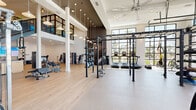 Located in Sanford, Florida, this expansive fitness center delivers a premium workout environment designed with both function and aesthetics in mind. The space spans a generously sized single floor, characterized by soaring double-height ceilings that create an open, airy atmosphere throughout the main exercise area. Large floor-to-ceiling black-framed windows and glass garage-style doors flood the interior with natural light while offering views of the surrounding outdoor landscape, including palm trees and the neighboring community.

The main workout floor features light hardwood-style flooring complemented by durable rubberized surfaces in key training zones. A dramatic central power rack and rig system anchors the free weights area, which includes a full selection of dumbbells, weight benches, and adjustable incline benches. Pendant lighting drops from the ceiling to add a stylish, modern touch alongside large ceiling fans that promote air circulation.

A dedicated cardio zone is equipped with treadmills, ellipticals, stair climbers, and stationary bikes, positioned to take advantage of the natural light and outdoor views. A separate, quieter room offers additional strength machines and benches in a more focused setting. A built-in storage and media station with a large display screen is conveniently located beneath an open staircase, adding a functional and connected touch to the overall facility.