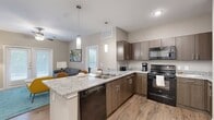 This 2-bedroom, 2-bathroom residence offers a thoughtfully designed single-floor layout that balances comfort and functionality. The open-concept kitchen is a standout feature, equipped with dark wood cabinetry, granite-style countertops, black stainless steel appliances including a refrigerator, range, microwave, and dishwasher, and a peninsula island with pendant lighting overhead. Light wood-style flooring flows throughout the kitchen and living areas, creating a cohesive, modern aesthetic. The living room is bright and welcoming, featuring a ceiling fan, large windows with blinds, and French doors that allow natural light to pour in. Both bedrooms are carpeted and well-sized, each with ceiling fans, double windows with outdoor views, and dedicated closets — one of which is a walk-in. The two bathrooms are finished with wood-toned vanities, light stone countertops, and wood-style flooring; one includes a shower/tub combination. A hallway connects the private areas of the home with ease. Neutral warm-beige tones throughout the space create a calm and inviting ambiance, making this a functional and comfortable place to call home.