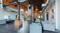 This single-floor space makes a bold architectural statement through its masterful blend of industrial heritage and modern design. The expansive lobby is anchored by two massive original brick columns — likely former boiler stacks — that rise dramatically toward the soaring exposed ceiling, where structural steel beams, wood decking, and visible HVAC ductwork reinforce the building's industrial origins. Large steel-framed windows along the street-facing wall flood the interior with abundant natural light while offering views of the surrounding neighborhood.

Dark slate tile flooring runs throughout, providing a sleek, durable foundation that complements the raw texture of white-painted brick walls. Contemporary light gray upholstered seating arrangements create comfortable gathering areas, balanced by warm orange accent walls that add energy and contrast. A beverage station with a coffee setup is tucked against one wall, adding a welcoming, functional touch.

A historic ornate metal door — preserved as an architectural artifact — serves as a striking focal point near the entrance corridor, which leads deeper into the building. Industrial-style pendant chandeliers provide ambient lighting, enhancing the space's character. The overall ambiance is one of adaptive reuse done thoughtfully, honoring the building's past while creating an inviting, contemporary environment.