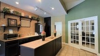 Located at 3851 Cape Landing Drive in Myrtle Beach, South Carolina, this single-story home offers a thoughtfully designed layout with a warm and inviting aesthetic throughout. The kitchen serves as a focal point of the home, showcasing striking black cabinetry paired with tile countertops and a spacious center island, ideal for meal preparation and casual dining. A herringbone-patterned tile backsplash in warm terracotta tones adds a distinctive decorative touch, complemented by sage green accent walls that bring a fresh, earthy palette to the space. Vaulted ceilings with track lighting enhance the sense of openness, while recessed lighting provides additional illumination. Black appliances maintain a cohesive, bold design throughout the kitchen. A French door visible from the kitchen hints at additional living spaces, offering an elegant transition between rooms. The tile flooring in neutral tones flows seamlessly through the space, providing both durability and continuity. Decorative vases and open shelving add personality and charm, making this home as functional as it is visually appealing. The overall ambiance is warm, stylish, and well-appointed.