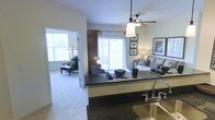 This single-story residence showcases an open-concept layout that connects the kitchen, living area, and an adjoining sitting room, creating a cohesive and functional flow throughout the space. The kitchen features dark granite countertops with a built-in sink, offering a sleek contrast against the light-colored walls and cabinetry. A decorative pendant light hangs above the kitchen island, complementing the ceiling fan installed in the living area, which provides both style and practicality. The living area is bathed in natural light, courtesy of large sliding glass doors that lead to an outdoor patio, enhancing the sense of openness. The space is furnished with a comfortable sofa accented by decorative pillows, alongside wall art featuring nature-inspired motifs. A doorway connects the main living space to an adjacent carpeted sitting room, which benefits from its own large window, allowing abundant natural light to filter in. This secondary room provides a quieter, more intimate setting. The overall ambiance of the space is bright, airy, and welcoming, with neutral tones throughout creating a clean, versatile aesthetic suitable for a variety of interior styles.