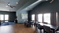 This single-floor space is designed to accommodate group gatherings and communal dining with a stylish, modern aesthetic. The room is defined by dramatic vaulted ceilings that rise to a prominent peak, adding a sense of openness and grandeur to the interior. Ceiling fans are mounted at varying heights to maintain airflow throughout the space.

The flooring is warm hardwood, which contrasts beautifully against the deep slate-gray walls that lend the space a contemporary, sophisticated character. Multiple dining table sets, each surrounded by upholstered chairs in neutral tones, are arranged throughout the room to comfortably seat several groups simultaneously.

A large, colorful abstract painting serves as a focal point on one wall, adding an artistic touch and a welcome pop of color against the dark backdrop. On the opposite side, a row of generously sized windows draped with elegant curtains allows abundant natural light to pour in, brightening the space and offering views of the surrounding exterior.

Recessed lighting is integrated into the ceiling to supplement natural light during evening hours. A media or entertainment setup is visible along one wall, suggesting the space may also serve as a venue for presentations or events. Overall, the room strikes a balance between comfort and functionality, making it well-suited for dining, socializing, or community events.