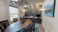 This business center combines functionality with refined design, offering residents or guests a productive and comfortable environment. The space is defined by soaring ceilings that contribute to an open, airy feel, while large windows fitted with roller shades allow ample natural light and views of the surrounding exterior.

At the center of the room, a substantial round wooden dining-style table surrounded by dark wood chairs provides a collaborative meeting or work area, anchored by a teal geometric-patterned area rug. Overhead, two striking black wrought-iron lantern-style pendant lights serve as prominent design focal points, lending an elegant yet contemporary character to the space.

Along one wall, a dedicated workstation features an extended granite-topped counter accommodating multiple computer stations and a shared printer, making it well-suited for productivity. The workstation wall is finished in a deeper taupe tone, with illuminated recessed niches displaying decorative teal ceramic vases, adding a curated aesthetic touch. Wall sconces provide additional ambient lighting throughout.

The entry features a modern paneled door alongside a glass-paneled door, contributing to the polished, upscale feel. Tile flooring, neutral warm-toned walls, and carefully selected décor complete the professional yet inviting atmosphere of this well-appointed business center.