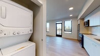 This single-floor unit showcases a clean, contemporary design characterized by light hardwood flooring, crisp white walls, and recessed lighting throughout. The open-concept layout connects the living area directly to the kitchen, creating a cohesive and functional living space. Large black-framed windows and a glass-paneled exterior door flood the interior with abundant natural light while offering views of surrounding trees and neighboring structures. A private balcony is accessible through the main living area door.

The kitchen is well-appointed with two-tone cabinetry—white lower cabinets paired with light wood upper cabinets—complemented by light-colored countertops and a white subway tile backsplash. Stainless steel appliances include a refrigerator, electric range, over-the-range microwave, and dishwasher.

A dedicated in-unit laundry closet houses a stacked washer and dryer. The bathroom features light hardwood flooring, a single-sink vanity with white cabinetry, a large mirror, and grab bars along the walls. A closet with open shelving provides additional storage. The unit's modern finishes and thoughtful layout create a bright, functional, and inviting living environment.