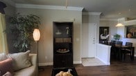 This single-story space presents a comfortable and cohesive interior defined by warm neutral wall tones and a thoughtful mix of flooring materials. The living area features rich hardwood-style flooring, while the entryway transitions to light-toned tile, creating a natural delineation between zones. Crown molding runs along the ceiling throughout, adding a refined architectural detail that elevates the overall aesthetic.

A dark espresso bookcase serves as a focal point in the living area, offering open shelving for decorative displays alongside enclosed cabinet storage below. Decorative accents, including woven baskets and small vases, lend a curated, lived-in warmth to the space. A softly glowing lantern-style floor lamp and a lush indoor plant contribute to the inviting ambiance.

The entryway is anchored by a clean white paneled door and benefits from an open sightline into the living space, enhancing the sense of flow. A mirrored surface near the entry reflects light and adds visual depth. Overall, the space conveys a relaxed yet polished atmosphere with consistent design details throughout.