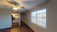 This efficiency unit in Houston, TX is a well-designed single-floor residence that maximizes functionality within a compact footprint. The open-concept layout seamlessly connects the living area to the kitchen, creating a cohesive and practical living space. Dark hardwood-style flooring runs throughout, providing a warm, consistent aesthetic that ties the rooms together.

The living area is bright and welcoming, featuring a ceiling fan with integrated lighting and large windows fitted with horizontal blinds that allow for ample natural light while maintaining privacy. Neutral gray walls create a clean, versatile backdrop throughout.

The kitchen is efficiently appointed with white cabinetry, dark granite-style countertops, a black refrigerator, a gas range with an over-the-range microwave, and a sink with overhead track lighting. A structural column defines the boundary between the kitchen and living space, adding architectural character.

The bathroom includes a full tub-shower combination with white tile surround, a toilet, and a vanity with matching dark countertop. Textured walls add a tactile design element consistent with the rest of the unit.

A textured popcorn ceiling is present throughout, and the unit includes climate control with visible ceiling vents. This efficient Houston residence offers a practical and cohesive living environment suited for individuals seeking a streamlined lifestyle.