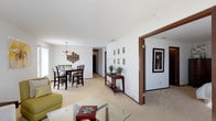 This well-designed open-concept living space offers a seamless blend of functionality and style. The area features a dining section with a contemporary high-top table and chairs, perfect for casual meals or entertaining. A striking geometric light fixture above the table adds a touch of modern elegance.

The room's neutral color scheme, dominated by white walls and light flooring, creates a bright and airy atmosphere. Textural interest is added through a textured accent wall in the dining area. Strategic pops of color, such as the lime green armchair visible in the foreground, enliven the space.

A glass coffee table in the living area adds a contemporary touch, while artwork on the walls provides visual interest. The open layout allows for easy movement between spaces, with wide doorways leading to adjacent rooms. A console table in the hallway offers both decorative and practical storage solutions.

Recessed lighting throughout ensures even illumination, complemented by natural light from unseen windows. The overall design aesthetic is clean and uncluttered, promoting a sense of spaciousness and calm in this inviting home environment.