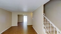This three-floor space offers a well-organized layout with a welcoming open flow between its main living areas. The main level features warm hardwood flooring that extends throughout, complemented by neutral beige walls and white baseboards that create a clean, cohesive aesthetic. A staircase with white railings is conveniently positioned along one wall, providing easy access to the upper levels, while carpeted steps add a practical touch.

The living and adjoining areas benefit from natural light streaming through vertical blinds, creating a bright and airy atmosphere. The textured ceilings add a subtle visual element to the otherwise understated interior. The open sightline between the main living area and the adjacent room enhances the sense of spaciousness, making the layout feel connected and functional.

The neutral color palette throughout provides a versatile backdrop suitable for a variety of interior styles. With three floors of living space, the property offers ample room for distinct living, sleeping, and utility zones, catering to a range of practical needs across its multiple levels.