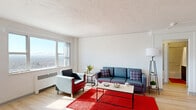This well-designed one-bedroom, one-bathroom unit offers a functional and comfortable layout across a single floor. Light hardwood flooring runs throughout, lending warmth and continuity to the space.

The living room is a standout area, bathed in natural light through large windows that frame sweeping panoramic city views. The open and airy atmosphere is complemented by a textured ceiling and a radiator heating unit positioned beneath the window.

The combined dining room and kitchen is both practical and inviting. The kitchen features dark espresso cabinetry, stainless steel appliances including a refrigerator, microwave, and electric range, and a clean tile backsplash. Adjacent to the kitchen, the dining nook is brightened by corner windows that pour in natural light, making it an ideal space for everyday meals.

The bedroom is a comfortable retreat, equipped with a wall-mounted air conditioning unit, a large window with city views, and a built-in closet. The hallways connecting the rooms provide efficient circulation throughout the unit.

The bathroom is clean and simply finished, featuring a full-sized tub and shower combination with white tile surround and a dark vanity cabinet. The overall ambiance of the unit is bright, clean, and modern, well-suited for those seeking a compact yet complete living space.