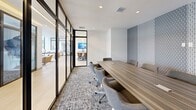 Located at 11265 Biscayne Blvd in Miami, FL, this sleek and contemporary conference room is designed to facilitate professional meetings in a polished, well-appointed environment. The room centers around a substantial wood-grain conference table that comfortably seats multiple participants in ergonomic upholstered chairs, creating a functional yet refined atmosphere. A wall-mounted large-format flat-screen television is positioned above a modern credenza — complete with gold-accented drawer pulls — making the space well-equipped for presentations and collaborative sessions. A striking geometric-patterned accent wall in muted blue and gray tones adds visual depth and a sophisticated design element to the room. The flooring features a textured, multi-tone carpet that complements the neutral and earthy color palette throughout. One full side of the room is enclosed by sleek, floor-to-ceiling black-framed glass partitions, providing a transparent boundary that connects the conference room visually to adjacent open workspaces, allowing natural light to filter through while maintaining acoustic separation. Recessed ceiling lighting ensures consistent, even illumination across the room. The overall design balances contemporary aesthetics with practical functionality, making it an ideal setting for professional gatherings in the heart of Miami.