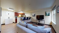 This single-floor unit offers a thoughtfully designed open-concept layout that seamlessly connects the main living areas. The living room is anchored by a comfortable sectional sofa, a glass coffee table, and a large flat-screen TV mounted on a sleek media console, with a window providing views of surrounding greenery and bringing in natural light.

The kitchen is efficiently designed with rich reddish-brown cabinetry, stainless steel appliances including a refrigerator and range, and a microwave. A peninsula with a warm wood-toned countertop extends into the living space, lined with modern black bar stools — ideal for casual dining or entertaining.

The primary bedroom features carpeted flooring, a full-size bed with clean white linens, and a walk-in closet with built-in shelving for ample storage. A second bedroom offers wood-look flooring, a window overlooking lush trees, a ladder shelf, and wall art that adds a cozy, personalized touch.

The bathroom is clean and functional, featuring light wood flooring, a long vanity with a gray countertop and generous cabinet storage, and a well-lit mirror. A dedicated utility and storage room with wire shelving and a water heater rounds out the space, providing practical organizational capacity throughout.