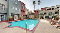 This outdoor communal area serves as the central gathering space of a multi-story residential complex, designed with both functionality and leisure in mind. At its heart lies a generously sized swimming pool with vivid turquoise water, framed by brick-edged coping and a wide concrete pool deck that provides ample space for relaxation and movement.

The surrounding buildings rise several stories and feature a warm color palette of terracotta reds and earthy tans, with stucco exteriors and multiple balconies overlooking the pool courtyard. Tall palm trees punctuate the space, lending a distinctly Southern California ambiance and adding vertical interest against the open sky.

The pool deck is well-equipped for outdoor living, featuring lounge chairs and patio umbrellas arranged along one side for shaded relaxation. A dedicated BBQ station provides an outdoor cooking area, ideal for community gatherings. A small utility structure near the pool houses additional amenities, while decorative potted plants — including a striking agave specimen in a terracotta urn — add greenery and character to the hardscaped surroundings.

Access points and gated areas are clearly integrated into the layout, ensuring a secure and organized environment. The overall design creates an inviting, resort-style atmosphere that encourages outdoor enjoyment within a well-maintained, community-oriented setting.