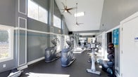 This fitness center is designed to support a complete workout experience within a bright, open, and well-appointed space. The room's most striking architectural feature is its soaring vaulted ceiling, which lends an expansive, airy feel and is complemented by two ceiling fans that help maintain comfortable airflow throughout. Large windows along the walls flood the interior with generous natural light, creating an energizing atmosphere for exercise.

The flooring consists of durable rubber matting, practical for both heavy equipment and high-impact activity. The walls are painted in a cool slate gray with crisp white trim accents, giving the space a clean and modern aesthetic. Full-length mirrors line one wall, visually expanding the room while allowing users to monitor their form during workouts.

The fitness center is outfitted with a variety of cardio machines, including ellipticals and cycling equipment, as well as a multi-station cable and strength machine. Free weights and balance equipment are also visible, offering options for a well-rounded training routine. A wall-mounted television provides entertainment during workouts. The overall ambiance is functional, modern, and inviting.