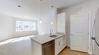 This single-floor studio showcases a thoughtfully designed open-concept layout that seamlessly integrates the kitchen and living areas. The kitchen serves as a central focal point, featuring a peninsula with light granite countertops, an undermount sink with a contemporary gooseneck faucet, and a stainless steel dishwasher. Crisp white cabinetry extends from base to upper units, providing ample storage, while tall cabinetry columns maximize vertical space. Two sleek pendant lights hang above the peninsula, adding a refined touch to the workspace.

The soaring ceilings amplify the sense of openness throughout the unit, creating an airy and expansive atmosphere. Light hardwood flooring runs continuously from the kitchen into the living area, unifying the space visually. A large window in the living area invites generous natural light, brightening the white walls and contributing to the clean, modern aesthetic. The overall design is minimalist and contemporary, emphasizing clean lines, neutral tones, and functional living within a compact yet well-appointed footprint.