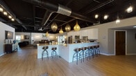 Located at 2219 Tanglewood Dr in Hammond, Indiana, this single-floor venue combines industrial architecture with sophisticated interior design to create a versatile and visually striking gathering space. The exposed ceiling structure, complete with dark-painted beams, wood slats, and visible ductwork, sets a dramatic backdrop throughout, while light hardwood flooring grounds the space with warmth.

The welcoming entry foyer features glass-panel doors that flood the space with natural light, pendant lighting, and seating areas that establish an inviting first impression. The open-concept main area transitions seamlessly between a full kitchen, bar seating, lounge, and dining zone. The kitchen is outfitted with stainless steel appliances, crisp white cabinetry, and light countertops, complemented by a sweeping bar peninsula lined with industrial-style wood and metal barstools. Eye-catching woven teardrop pendant lights hang above, adding an artisan touch.

The lounge area offers ample seating arranged around a sleek linear fireplace, with a bold gold chandelier serving as a focal point. Natural light pours in through sliding glass doors that open to an outdoor patio. The formal dining area accommodates a large party, anchored by geometric gold pendant fixtures and surrounded by windows offering serene outdoor views. Together, these spaces create a cohesive, elegantly appointed venue ideal for events and gatherings.