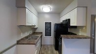 This single-floor kitchen is designed with a functional galley-style layout, maximizing the use of available space through parallel runs of cabinetry and countertops on both sides. White shaker-style upper and lower cabinets line the walls, providing ample storage while contributing a clean, bright aesthetic. The dark laminate countertops offer a striking contrast against the white cabinetry, creating a balanced visual appeal throughout the space.

On one side, a stainless steel double-basin sink is positioned beneath the cabinetry, accompanied by a dishwasher. The opposite side features black appliances, including a refrigerator, over-the-range microwave, and a matching black range, creating a cohesive color scheme. Wood-style plank flooring runs the length of the kitchen, adding warmth and durability underfoot.

A flush-mount ceiling light provides central illumination, complementing the textured ceiling finish. A framed chalkboard panel on the end wall serves as a charming decorative and functional accent, adding a personalized touch to the space. Overall, the kitchen balances practicality with a clean, modern aesthetic.