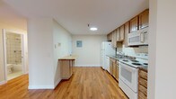 This single-floor unit is designed with a straightforward, functional layout that makes efficient use of its space. Warm hardwood flooring runs throughout, lending a cohesive and inviting feel to each area.

The kitchen is well-equipped with medium-toned wood cabinetry providing ample storage above and below the countertops. White appliances include a refrigerator, electric range, built-in microwave, and dishwasher. A granite-style sink with a chrome faucet is positioned along the counter run. A built-in breakfast bar with a butcher block surface extends from the cabinetry, offering a casual dining or prep area. An electrical panel is accessible on the adjacent wall, and a doorway connects to the rest of the unit.

The living area is open and bright, with a sliding glass door fitted with vertical blinds that allows natural light to fill the room. A wall-mounted air conditioning unit and a baseboard heating system ensure year-round comfort. The neutral white walls throughout create a clean, versatile backdrop suitable for a variety of interior styles. A bathroom with tile walls is also accessible within the unit.