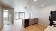 Located at 4929 Shelby Ave in Rapid City, South Dakota, this single-level unit offers a thoughtfully designed layout with a clean, modern aesthetic throughout. The open-concept kitchen and living area serves as the heart of the space, anchored by a central island equipped with a built-in sink, dishwasher, and generous counter space finished in light-toned laminate. Gray cabinetry pairs with black appliances — including a refrigerator, electric range, and over-the-range microwave — to create a cohesive, contemporary kitchen. Pendant lighting above the island and recessed ceiling lights provide ample illumination across the open area. Light wood-look flooring flows seamlessly through the kitchen and living spaces, contributing to a bright, airy atmosphere. The living area features large windows and a set of glass-paned exterior doors that invite natural light and provide outdoor access. Carpeted bedrooms offer a softer, quieter retreat, with one featuring a closet with dedicated storage. The bathroom includes a stone-topped vanity and tile-style flooring for a clean, functional finish. A dedicated laundry area with an in-unit washer and dryer adds everyday convenience. The space is efficiently planned, making it well-suited for comfortable, low-maintenance living.