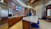 This thoughtfully designed single-floor space blends functionality with striking visual character. The centerpiece is a curved kitchen area adorned with an eye-catching multicolored mosaic tile feature wall that curves along the interior, creating a bold and lively focal point. Warm-toned wood cabinetry lines the kitchen, complemented by stainless steel appliances including a large French-door refrigerator, double wall ovens, and a dishwasher. A generously sized white quartz island with a rounded edge provides additional workspace and casual seating with vibrant blue-cushioned bar stools that echo the mosaic's colorful palette. A wall-mounted flat-screen television is prominently positioned above the counter, adding a media element to the kitchen environment. The flooring throughout is natural travertine stone in warm golden tones, lending an upscale, timeless quality underfoot. Adjacent to the kitchen, a welcoming entry corridor features exposed wood beam accents, arched doorways, rich dark wood trim, and a distinctive sculptural element near the entrance. Multiple intimate dining nooks are arranged throughout, each furnished with round wooden tables and blue-cushioned chairs. Large shuttered windows flood these seating areas with abundant natural light while offering views to the outdoors. Contemporary artwork adorns the walls, enhancing the refined yet approachable ambiance of the overall space.