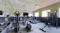 This fitness center is thoughtfully designed to inspire and motivate. The space is defined by its bold two-tone walls — a charcoal gray lower half contrasted with a vibrant yellow-green upper half — adorned with motivational fitness-themed typography including words like "determination," "focus," "motivation," and "discipline," creating an energizing atmosphere throughout.

The room is well-lit by a combination of overhead fluorescent panel lighting and natural light streaming in through multiple windows along one wall, giving the space a bright, open feel. A ceiling fan contributes to air circulation and comfort during workouts.

The fitness center is outfitted with a solid selection of cardio equipment, including multiple treadmills lined along the perimeter, upright and recumbent stationary bikes, and an elliptical cross-trainer, catering to a range of cardio preferences. A wall-mounted flat-screen television is positioned for easy viewing during exercise sessions.

The flooring consists of durable, low-pile carpet tiles suited for a high-traffic fitness environment. The layout is practical and spacious, allowing for comfortable movement between machines. Overall, this fitness center delivers a functional, well-appointed space with an atmosphere designed to keep users focused and driven.