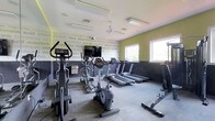 This fitness center is thoughtfully designed to inspire and motivate. The space is defined by its bold two-tone walls — a charcoal gray lower half contrasted with a vibrant yellow-green upper half — adorned with motivational fitness-themed typography including words like "determination," "focus," "motivation," and "discipline," creating an energizing atmosphere throughout.

The room is well-lit by a combination of overhead fluorescent panel lighting and natural light streaming in through multiple windows along one wall, giving the space a bright, open feel. A ceiling fan contributes to air circulation and comfort during workouts.

The fitness center is outfitted with a solid selection of cardio equipment, including multiple treadmills lined along the perimeter, upright and recumbent stationary bikes, and an elliptical cross-trainer, catering to a range of cardio preferences. A wall-mounted flat-screen television is positioned for easy viewing during exercise sessions.

The flooring consists of durable, low-pile carpet tiles suited for a high-traffic fitness environment. The layout is practical and spacious, allowing for comfortable movement between machines. Overall, this fitness center delivers a functional, well-appointed space with an atmosphere designed to keep users focused and driven.