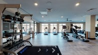 This two-floor fitness facility at 9 Tower Ln in New Haven, Connecticut, offers a comprehensive and well-appointed workout environment designed to support a wide range of fitness goals. The upper level is bathed in natural light, with large floor-to-ceiling windows lining the walls and providing views of the surrounding neighborhood. This bright, airy space features light wood-style flooring and houses a generous array of cardio equipment, including multiple treadmills, ellipticals, and cycling machines. A ceiling fan and recessed lighting keep the environment comfortable throughout. A sleek cable railing staircase connects the two floors, creating an open and modern transition between levels. The lower level takes on a more dynamic character, with bold teal-accented tile flooring and a striking orange accent wall. This floor is dedicated to strength training, featuring multi-functional cable machines, a full dumbbell rack, benches, and ample open floor space for functional training. Large mirrors are strategically placed throughout to maximize the sense of space. A dedicated studio area includes a large digital display wall with an on-demand fitness platform and medicine balls for group or guided workouts. A standout feature of the facility is the salt therapy room, which offers a tranquil retreat with glowing backlit Himalayan salt panels, reclining lounge chairs, and warm ambient lighting—ideal for post-workout recovery.