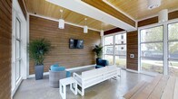 This contemporary single-floor space makes a strong architectural impression through its thoughtful combination of natural and modern materials. The walls are clad in rich, textured wood paneling that runs from floor to ceiling, complemented by a warm, exposed wood-slat ceiling accented by white structural beams, creating a cohesive and inviting aesthetic throughout. Polished concrete floors provide a sleek, durable foundation that contrasts beautifully with the warmth of the wood surfaces above.

The lounge area is anchored by a clean-lined white sofa and matching coffee tables, paired with stylish blue accent chairs and a rounded wicker seating piece, offering a comfortable and functional gathering space. Pendant cylinder light fixtures hang from the ceiling, providing focused ambient lighting that enhances the room's modern character.

A wall-mounted flat-screen television serves as a central focal point on the wood-paneled wall, making the space well-suited for both casual relaxation and social interaction. Lush tropical potted plants are strategically placed throughout, adding vibrant greenery that softens the architectural elements and enlivens the atmosphere.

Large floor-to-ceiling windows flood the interior with natural light and offer views of the surrounding exterior, seamlessly connecting the indoor environment with the outside world.